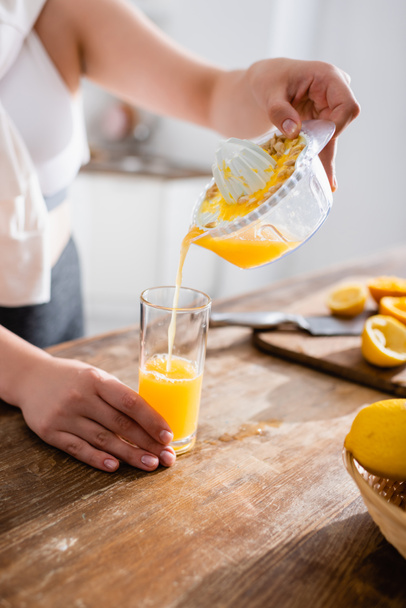 絞りたてのオレンジジュースを絞りたての果物の近くのガラスに注ぐ女性のクロップドビュー  - 写真・画像