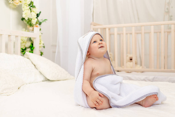 baby boy 8 months old sitting on a white bed in a white towel after bathing in the bathroom at home, place for text - Foto, imagen