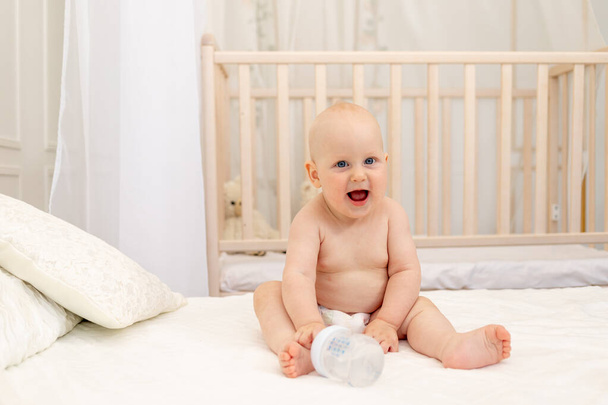 8 hónapos kisfiú pelenkában ül egy fehér ágyon egy üveg tejjel otthon, bébiétel koncepció, első csali, szöveg helye - Fotó, kép