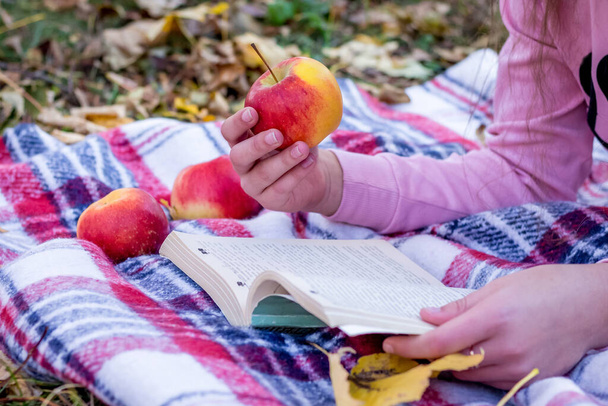 少女はリンゴを手に取って本を読む。秋の自然の中で休む - 写真・画像