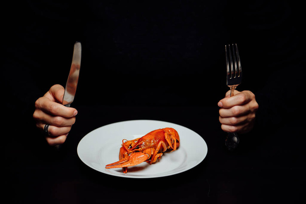 boiled crayfish on a white plate, knife and fork in hand on a black background minimalism - Fotografie, Obrázek
