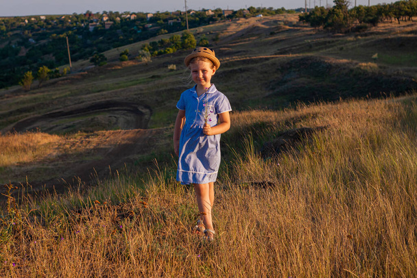 Чарівна маленька дівчинка в солом'яному капелюсі і синя літня сукня в трав'яному полі з дикими квітами в руках. Щаслива дитина з довгим світлим плетеним волоссям сільський пейзаж. смішна ходьба дитина на відкритому повітрі
 - Фото, зображення