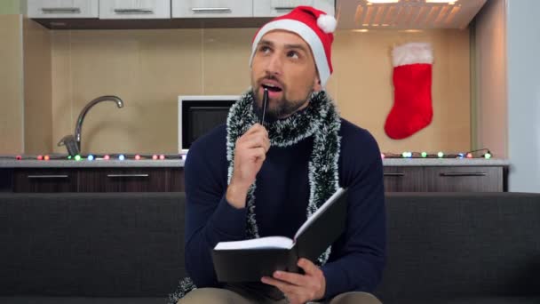 Man New Year Santa Claus hat thinks dreams next year, writes in notebook, smiles - Footage, Video
