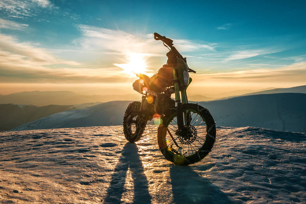 una motocicleta con cadenas sobre ruedas se encuentra en la cima de una montaña en invierno al atardecer - Foto, imagen