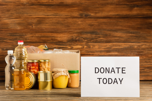 cardboard box with groats near water, oil, canned food, honey and donate today card on wooden background, charity concept - Photo, Image