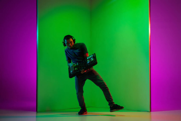 Young caucasian musician in headphones performing on bicolored green-purple background in neon light - Fotografie, Obrázek
