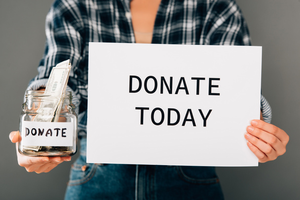 cropped view of woman holding card with donate today lettering and jar with money on grey background - 写真・画像