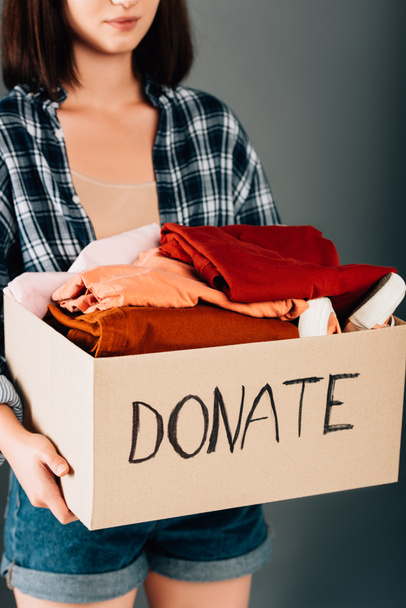 cropped view of woman holding box with donate lettering and clothes on grey background - Photo, Image