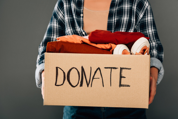 cropped view of woman holding box with donate lettering and clothes on grey background - Фото, зображення
