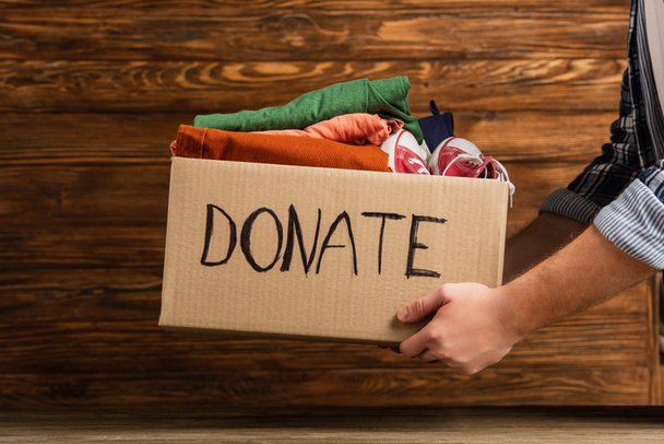 cropped view of man holding cardboard box with donate lettering and clothes on wooden background, charity concept - Photo, Image