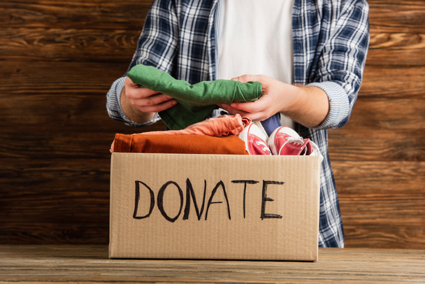 cropped view of man putting clothes in cardboard box with donate lettering on wooden background, charity concept - Foto, immagini