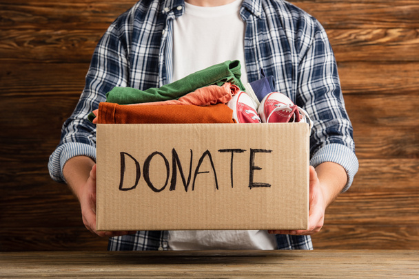 cropped view of man holding cardboard box with donate lettering and clothes on wooden background, charity concept - Foto, imagen