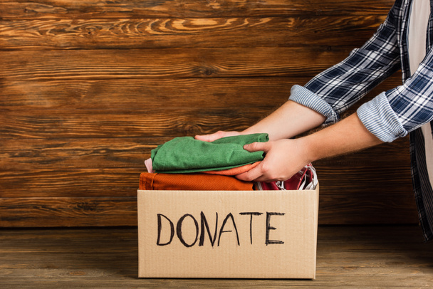 cropped view of man putting clothes in cardboard box with donate lettering on wooden background, charity concept - Фото, изображение