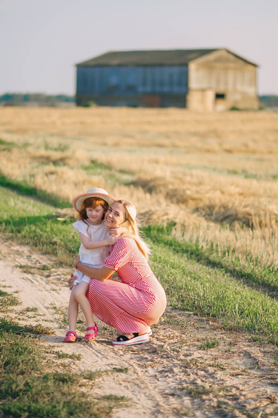 Η νεαρή μητέρα και η κοκκινομάλλα κόρη της με το ψάθινο καπέλο κάθονται στον ήλιο κοντά στη φάρμα στο λιβάδι μια φωτεινή καλοκαιρινή μέρα.. - Φωτογραφία, εικόνα
