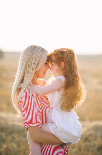 Retrato emocional de una joven madre rubia feliz y activa sosteniendo a su pequeña hija de heredita roja positiva en sus brazos mientras está de pie en un campo a la luz del atardecer. Vida de verano - Foto, Imagen