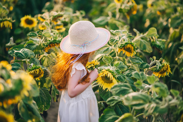 Cute little red-heired dziewczyna w słomkowym kapeluszu ze słonecznikiem w lecie pole słonecznika na zachodzie słońca. Koncepcja szczęścia dzieci. Widok z bliska - Zdjęcie, obraz