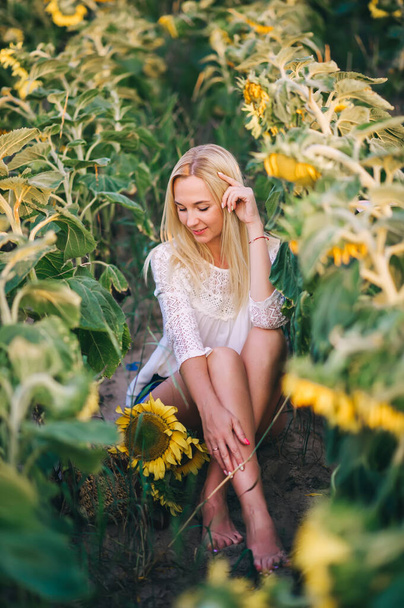 Portret młodej blondynki w białej bluzce siedzącej na tle słoneczników. Zbliżenie. - Zdjęcie, obraz