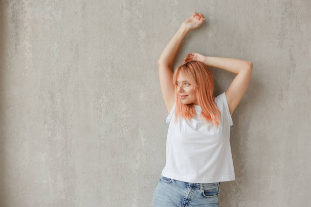 Giovane donna gioiosa in t-shirt bianca con i capelli tinti posa a parete con le mani alzate - Foto, immagini