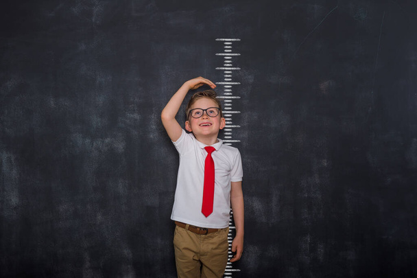 Little kid boy measuring himself. School boy measuring his growth in height against a blackboard scale. Back to school. Pupil in uniform - Photo, Image