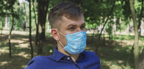 A guy in a medical mask sits on a wooden bench in the park - Photo, Image