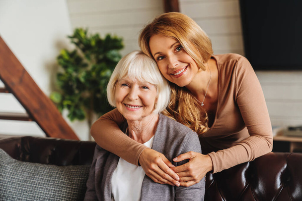 Cheerful middle aged woman embracing senior mother at home and looking at camera - Fotoğraf, Görsel