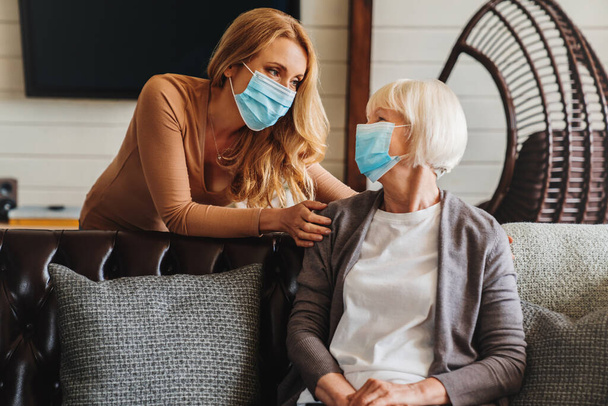 Femme âgée avec masque médical et assistante sociale lui rendant visite à la maison - Photo, image