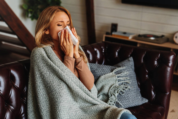 Sick woman covered with blanket sneezing in napkin while sitting on sofa - Foto, imagen