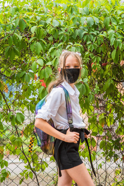 Schoolmeisje meisje met een zwart beschermend masker met een rugzak. Schoolmeisje in een wit shirt en zwarte broek, schooluniform. Vallen. Binnenkort terug naar school.. - Foto, afbeelding