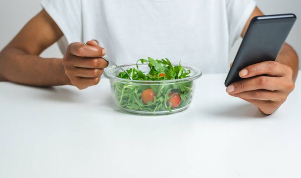 τα χέρια μιας μαύρης Αφρο-γυναίκας που τρώει σαλάτα από ένα κουτί φαγητού ενώ χρησιμοποιεί ένα έξυπνο τηλέφωνο. επιλεκτική εστίαση στη σαλάτα - Φωτογραφία, εικόνα