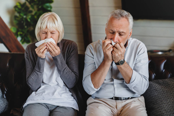 Virus and flu concept. Mature couple sitting on sofa and blowing their noses. - Photo, image
