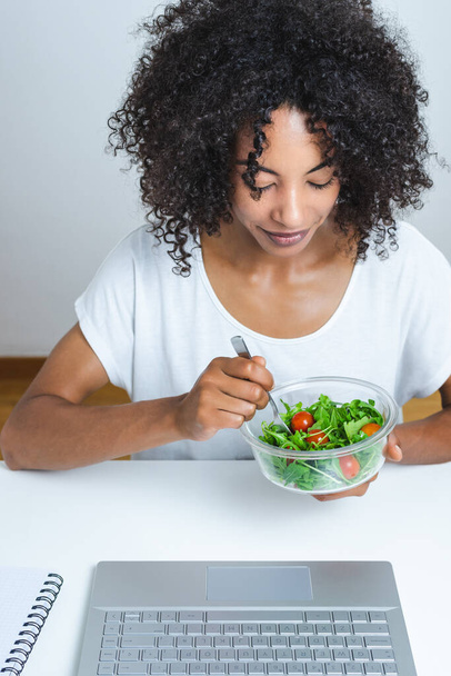 mooie jonge zwarte vrouw eten een salade in de voorkant van een moderne laptop met witte achtergrond - Foto, afbeelding