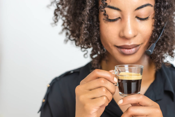 xícara transparente com café espresso cremoso preto segurar por uma bela mulher negra afro com um fone de ouvido helpdesk. foco seletivo no copo
 - Foto, Imagem
