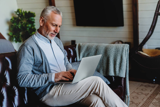 Handsome old man using laptop and smiling while sitting on couch at home - Fotoğraf, Görsel