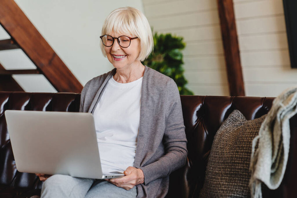 Senior woman in living room with laptop smiling while sitting on sofa - Photo, image