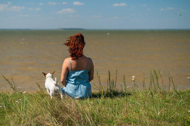 Rear view of a sitting woman on the river bank with a dog. Girls spend weekends in nature with a pet. - Foto, afbeelding