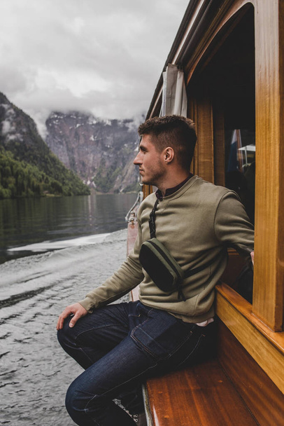 Brunet kaukasischen Mann sitzt auf dem Fenster des hölzernen Bootes auf dem Hintergrund des Sees, Wald und Felsen Berg in Wolken. Königssee. Bayern. Deutschland - Foto, Bild