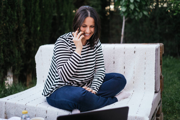 Брюнетка, разговаривающая по мобильному телефону, улыбается в саду своего дома, когда телепортируется со своим ноутбуком. На ней темные джинсы и синий и белый полосатый свитер. Она сидит на диване из деревянных поддонов. Сад полон деревьев - Фото, изображение