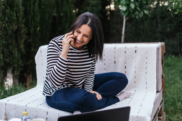 Brunette woman talking on the cellphone smiles in the garden of her house while teleworking with her laptop. She is wearing dark jeans and a blue and white striped sweater. She is sitting in a couch made of wooden pallets. The garden is full of trees - Photo, Image