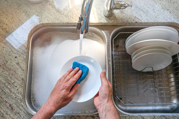 A man washes a white plate with a blue foam sponge in the kitchen, top view. Dishwashing, home routine, cleaning up the plates. - Foto, Imagem