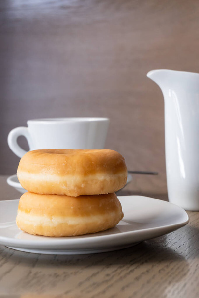 Due ciambelle fresche per la colazione su un tavolo di legno, una tazza di caffè espresso e una brocca di latte - Foto, immagini