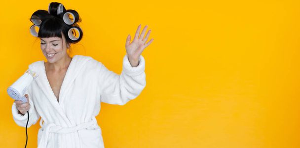 young beautiful woman housewife in a white robe. curlers on the head. Yellow background. Glove-shaped washcloth. hold the hair dryer in your hands and sing songs - Photo, Image