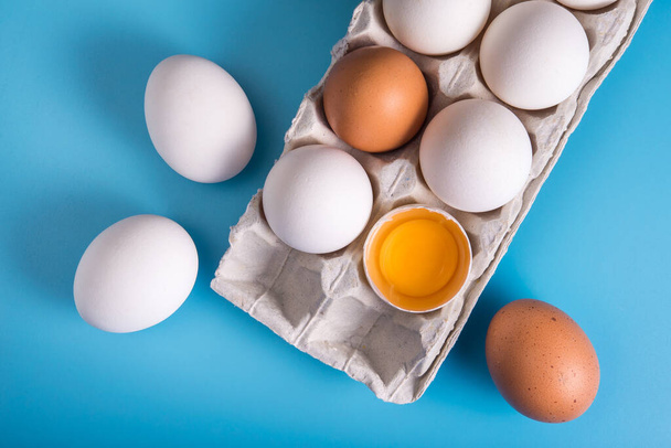 Homemade chicken eggs in a tray. One is broken. Blue background. Top view - Photo, Image