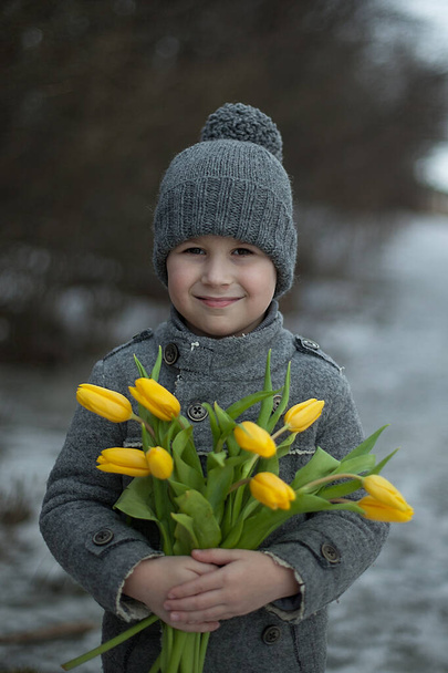 Przystojny chłopiec w szarym płaszczu gratuluje wakacji dużymi bukietami żółtych tulipanów. 8 marca, Dzień matki, Dzień Kobiet, Dar, niespodzianka, emocje. - Zdjęcie, obraz