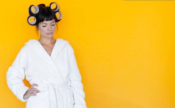 young beautiful woman housewife in a white robe. curlers on the head. Yellow background. put your face down and think. - Photo, Image