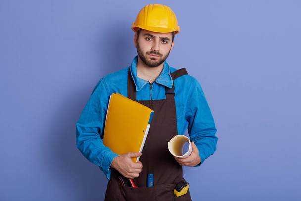 Young serious engineer with paper folders in hands, looking directly at camera with confident facial expression, wearing helmet and apron, stands against blue wall. - Foto, Imagem