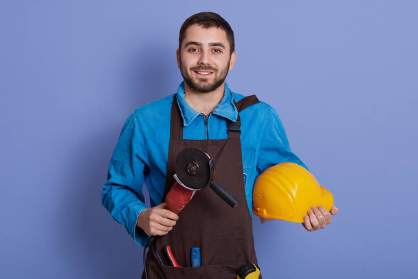 Smiling bearded man wearing brown apron holding grinder tool and yellow protective helmet in hands isolated over blue background, looks smiling at camera. - Фото, зображення
