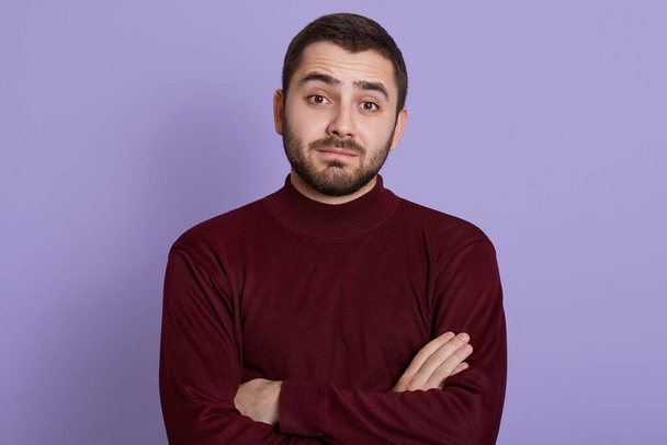 Thoughtful young man with skeptic, doubtful, distrustful look posing against lilac background with folded hands, wearing burgundy sweater, bearded puzzled male. - Photo, Image