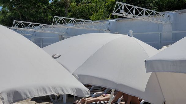 Personas irreconocibles toman el sol bajo sombrillas blancas en la playa. Sombras de sol de playa en Europa. Escena de verano - Foto, imagen