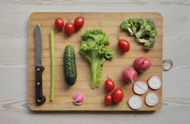 Fresh vegetables on a cutting board on a light wooden table Tomatoes, cucumber, lettuce, broccoli, radish, knife - Photo, image