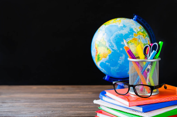 Back to school. Stack of books, school supplies, globe and glasses on black board background. Education concept. Copy space. Banner - Photo, Image